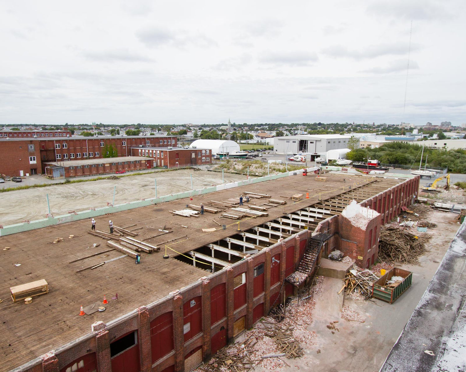 View of old brick mill building with the third floor ripped off and the 2nd floor exposed. The photo is taken from on top of the roof of the building next to it. You can see brick rubble and yellow pine boards being stacked by demolition workers.