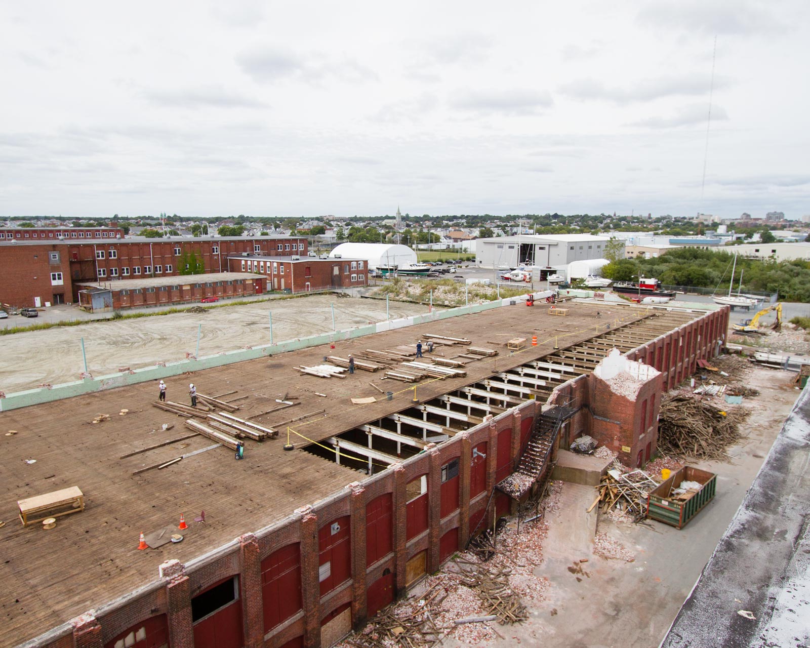 View of old brick mill building with the third floor ripped off and the 2nd floor exposed. The photo is taken from on top of the roof of the building next to it. You can see brick rubble and yellow pine boards being stacked by demolition workers.